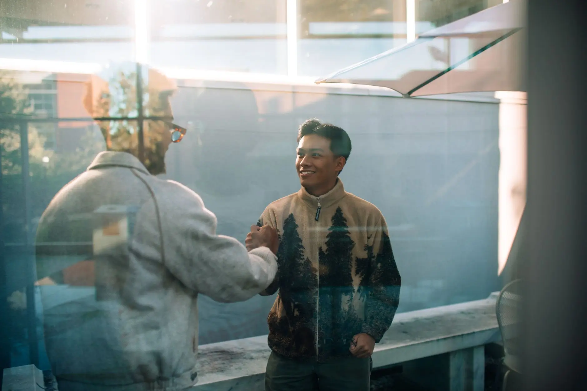 Two men smile and shake hands through a glass window, reflecting a warm friendly interaction.